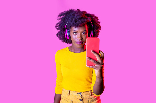 Young black woman holding smartphone taking selfie or videcalling wearing wireless headphones isolated background - Foto, Bild