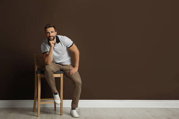 Handsome man sitting on stool near brown wall. Space for text - Zdjęcie, obraz
