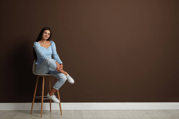 Beautiful young woman sitting on stool near brown wall. Space for text - Fotografie, Obrázek