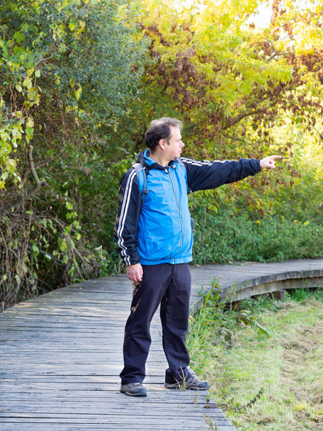 Man pointing away, middle age tourist enjoying walk in autumn nature, wooden path, sunny day - Photo, Image