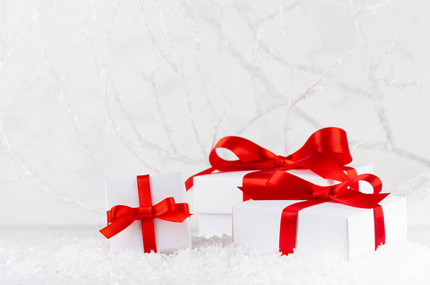 White christmas festive background - gift boxes with satin red ribbon and bow in decorative snow and soft light frosty winter forest with white branches, copy space. Holiday presents for celebration. - Photo, image