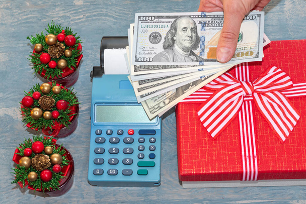 A hand holding dollars over a cash register near a red gift box with a ribbon and small Christmas trees with cones - Foto, immagini