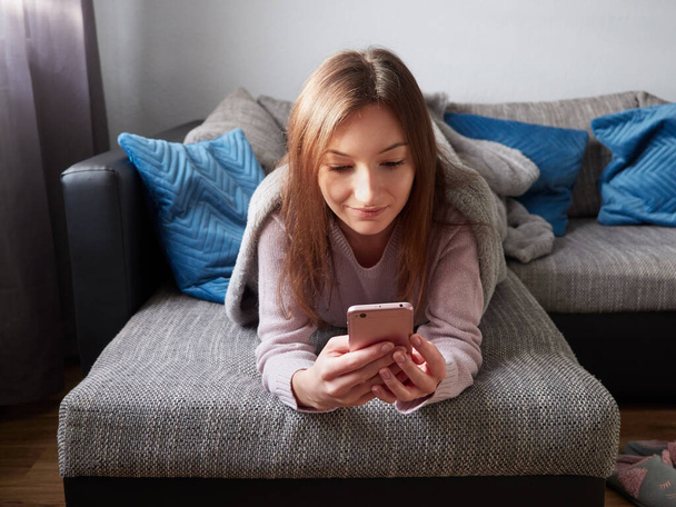 Młoda, szczęśliwa kobieta leży na kanapie pokryta kocem i używa smartfona. Atrakcyjna dziewczyna w salonie przegląda internet na smartfonie. - Zdjęcie, obraz