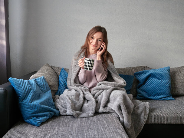 Молодая, счастливая женщина сидит на диване, покрытом одеялом. Улыбающаяся девушка разговаривает по телефону и держит кружку с горячим напитком в руке. - Фото, изображение