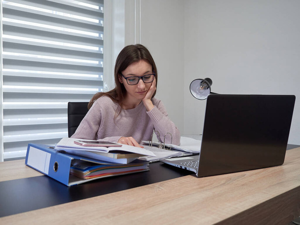 Mladá žena s brýlemi pracuje doma a používá notebook. Dívka sedí u stolu a rukou si opře hlavu. Na stole jsou pořadače s dokumenty. Domovská kancelář - Fotografie, Obrázek