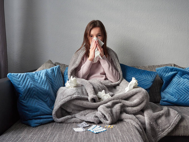Young sick girl sitting on a couch under a blanket. A woman with a runny nose is blowing her nose into a handkerchief. There are various packets of tablets in front of her. There are used handkerchiefs scattered on the couch. - 写真・画像