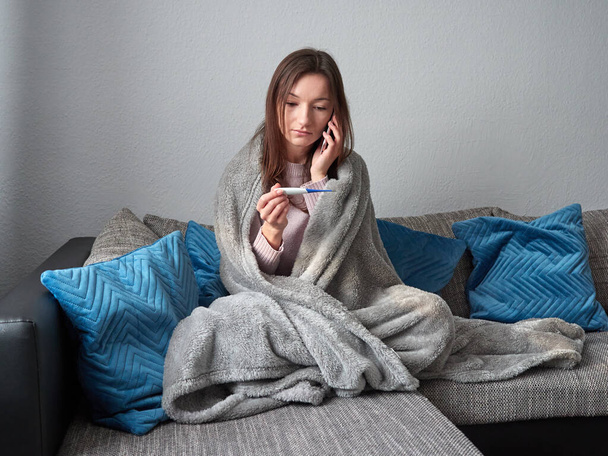Young sick girl sitting on a couch under a blanket. The woman checks her body temperature. She's calling the doctor. Telephone consultation - Fotoğraf, Görsel