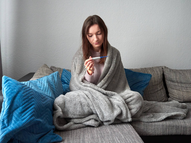 Νεαρό άρρωστο κορίτσι κάθεται σε έναν καναπέ κάτω από μια κουβέρτα. Η γυναίκα ελέγχει τη θερμοκρασία του σώματός της.. - Φωτογραφία, εικόνα