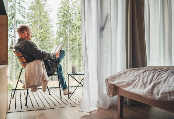 Alone man sitting in chair on country house balcony and enjoying forest view, tea cup, fresh air and bestseller novel thick book. Cozy bedroom interior back view. Reading or education concept image - Photo, image