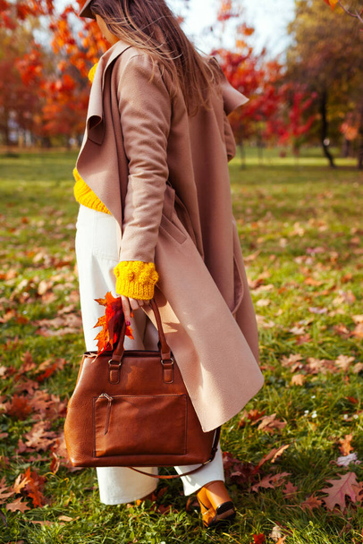 Fashionable woman wearing stylish clothes and accessories walking in fall park. Autumn female outfit. Trendy flared pants with yellow sweater, beige coat and brown handbag - Foto, Bild
