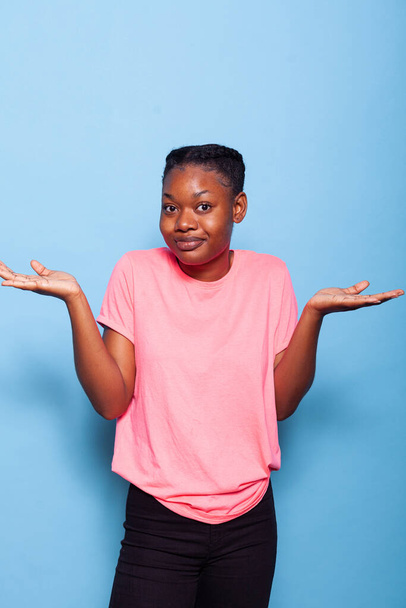 Портрет смущенной африканской молодой женщины в розовой футболке на синем фоне - Фото, изображение