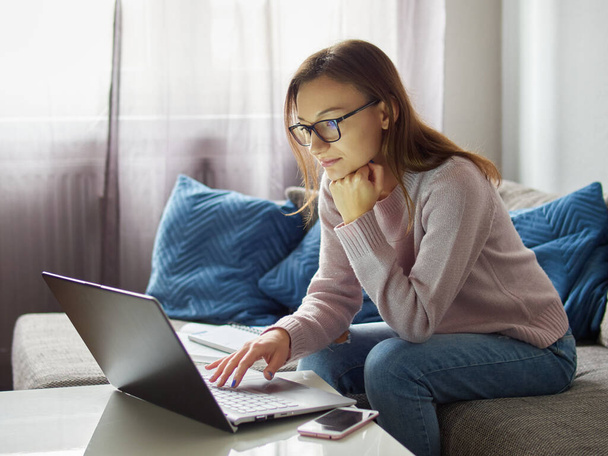 Giovane donna con gli occhiali seduta sul divano e che lavora su un computer portatile. Una studentessa sostiene la testa con la mano. Ragazza nel soggiorno di fronte al computer - Foto, immagini