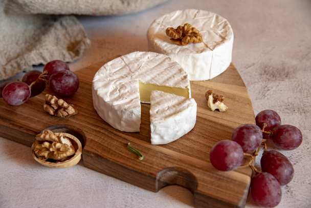 Cheese with white mold, grapes and walnuts on a wooden cutting board on a light countertop. - Valokuva, kuva