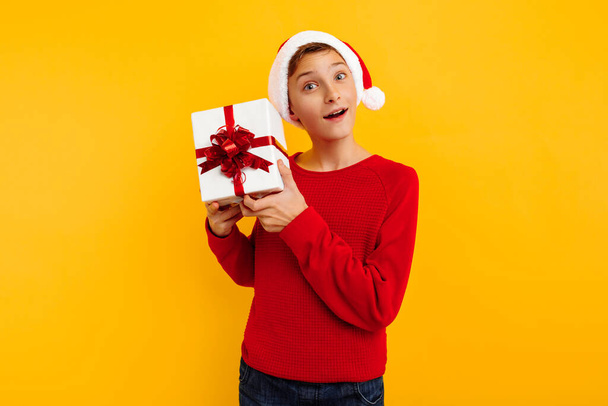Pensive boy in a Christmas hat and a gift in his hands. Thoughtfully thinks what is inside the gifts. On a yellow background. Christmas concept - Fotografie, Obrázek