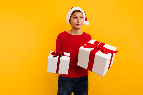 Pensive boy in a Christmas hat and a gift in his hands. Thoughtfully thinks what is inside the gifts. On a yellow background. Christmas concept - 写真・画像
