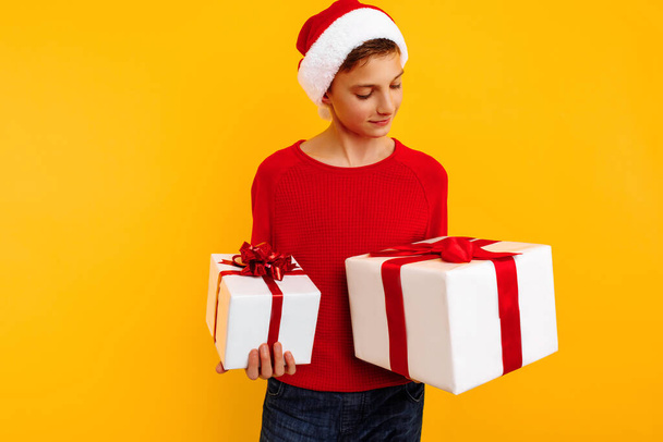 Pensive boy in a Christmas hat and a gift in his hands. Thoughtfully thinks what is inside the gifts. On a yellow background. Christmas concept - Photo, image