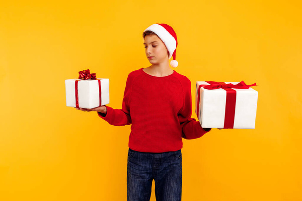 Pensive boy in a Christmas hat and a gift in his hands. Thoughtfully thinks what is inside the gifts. On a yellow background. Christmas concept - Foto, Imagen