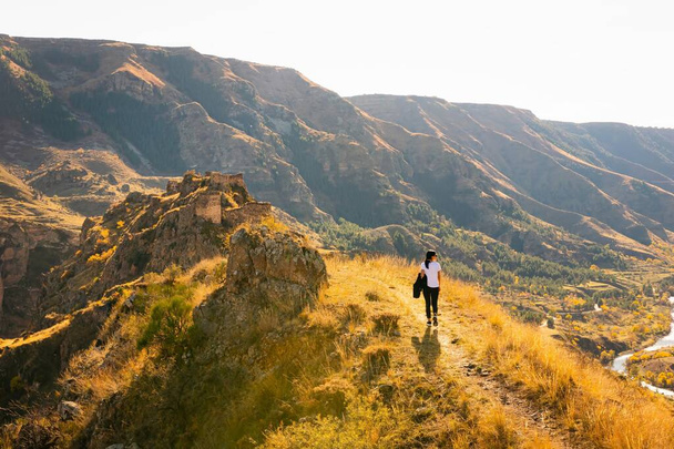 Femme monte la colline vers la forteresse de Tmogvi entourée d'une nature d'automne dorée et d'un paysage spectaculaire dans le caucase. Automne chaud couleurs dorées nature à l'extérieur - Photo, image