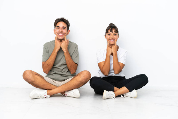 Young mixed race couple sitting on the floor isolated on white background smiling with a happy and pleasant expression - Photo, Image