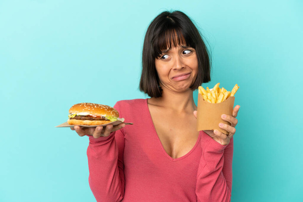 Nuori brunette nainen tilalla hampurilainen ja paistettuja sipsejä yli eristetty sininen tausta - Valokuva, kuva