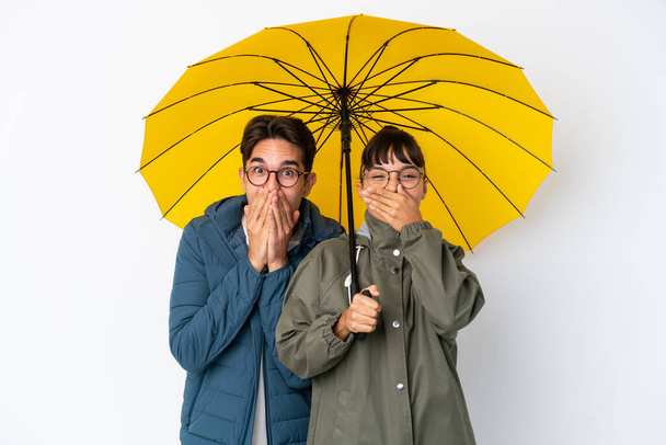 Joven pareja de raza mixta sosteniendo un paraguas aislado sobre fondo blanco sonriendo mucho mientras cubre la boca - Foto, imagen