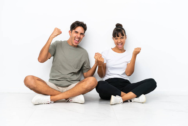 Young mixed race couple sitting on the floor isolated on white background celebrating a victory - Photo, Image
