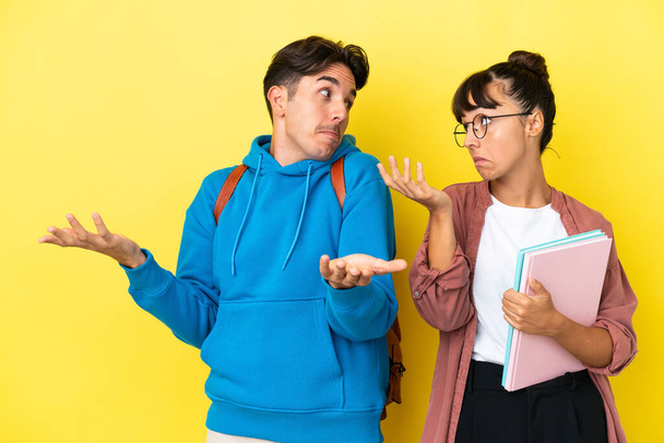 Young student couple isolated on yellow background making unimportant gesture while lifting the shoulders - Photo, Image