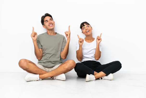 Junges gemischtes Paar, das isoliert auf weißem Hintergrund auf dem Boden sitzt und mit dem Zeigefinger auf eine großartige Idee zeigt - Foto, Bild