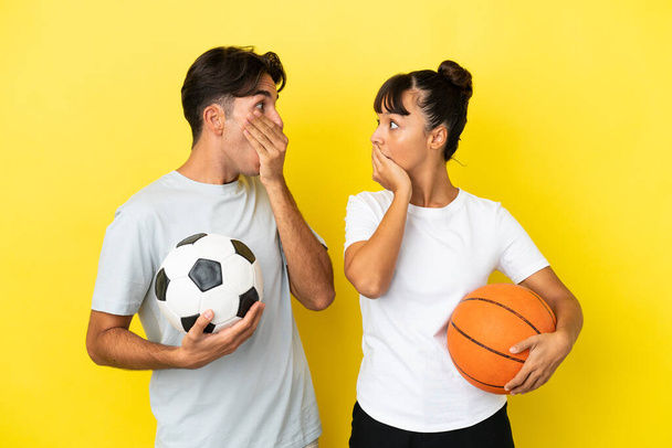 Futbol ve basketbol oynayan genç çift, sarı arka planda izole edilmiş ağızlarını uygunsuz bir şey söyledikleri için elleriyle kapatıyor. - Fotoğraf, Görsel
