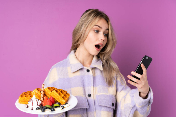 Teenager Russian girl holding waffles isolated on purple background holding coffee to take away and a mobile - 写真・画像