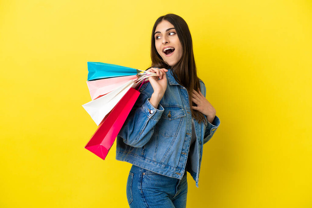 Young caucasian woman isolated on blue background holding shopping bags and smiling - Foto, afbeelding