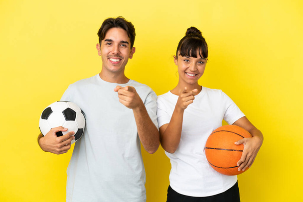 joven deporte pareja jugando fútbol y baloncesto aislado en amarillo fondo puntos dedo en usted con una expresión de confianza - Foto, imagen