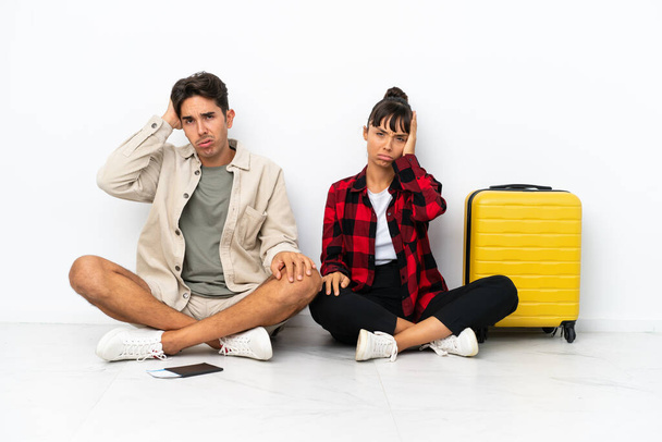 Jeune couple mixte de voyageurs assis sur le sol isolé sur fond blanc avec une expression de frustration et de ne pas comprendre - Photo, image