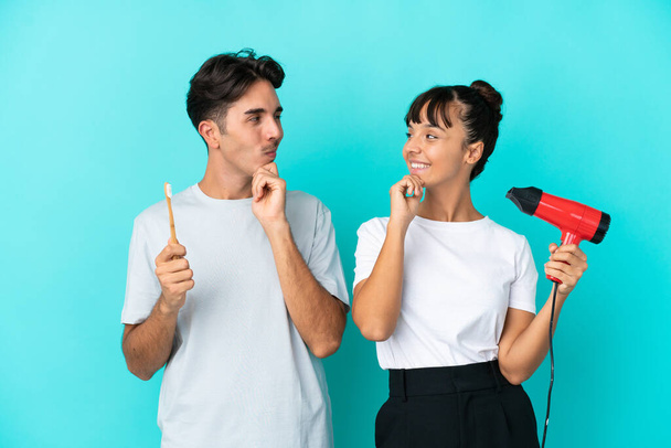 Young mixed race couple holding a hairdryer and toothbrush isolated on blue background looking looking at each other - Photo, Image