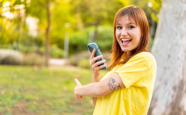 Молода красива руда жінка на відкритому повітрі, використовуючи мобільний телефон і вказуючи назад
 - Фото, зображення
