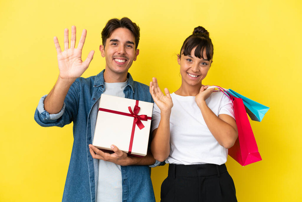 Young couple holding shopping bags and present isolated on yellow background saluting with hand with happy expression - Photo, Image