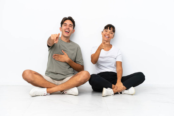 Nuori sekarotuinen pari istuu lattialla eristyksissä valkoisella taustalla osoittaen sormella jotakuta ja nauraen paljon - Valokuva, kuva
