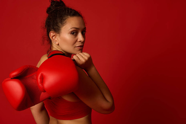 Attractive wavy haired pretty female athlete, woman boxer fighter poses against colored background with red boxing gloves looking confidently at camera through her shoulder. Martial art concept - Photo, Image