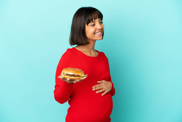 Mujer embarazada joven sosteniendo una hamburguesa sobre fondo aislado mirando lado - Foto, Imagen