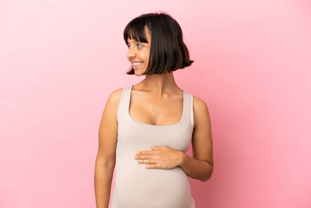 Junge schwangere Frau über isoliertem rosa Hintergrund, der zur Seite schaut und lächelt - Foto, Bild