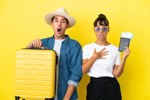 Jóvenes amigos viajeros sosteniendo una maleta y pasaporte aislados sobre fondo amarillo con expresión facial sorpresa y conmocionada - Foto, Imagen