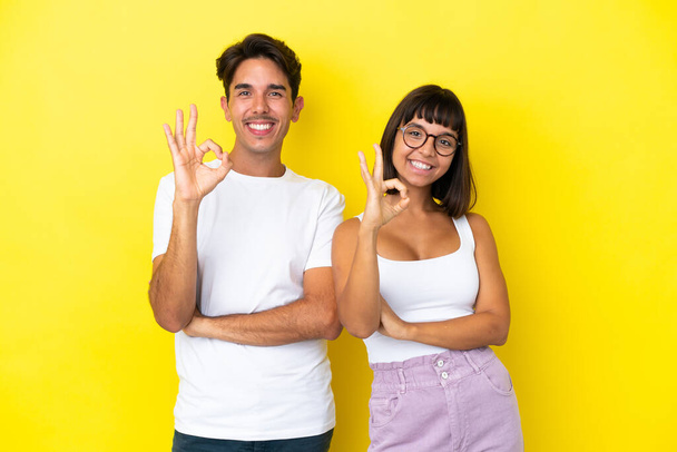 Junges Mischlingspaar isoliert auf gelbem Hintergrund und zeigt ein Ok-Zeichen mit Fingern - Foto, Bild