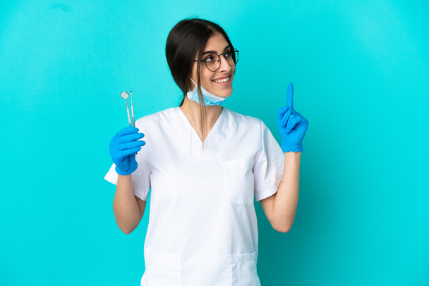 Joven dentista caucásica mujer sosteniendo herramientas aisladas sobre fondo azul señalando una gran idea - Foto, Imagen