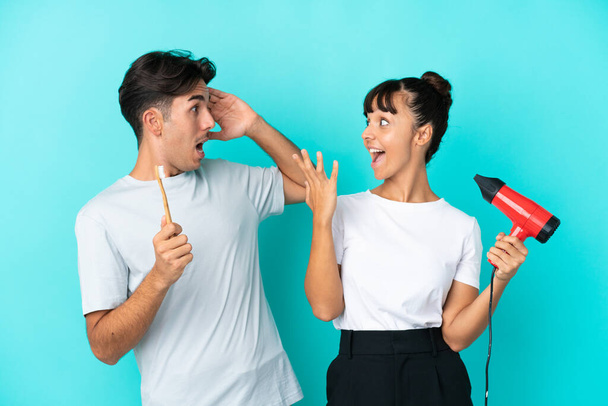 Young mixed race couple holding a hairdryer and toothbrush isolated on blue background with surprise and shocked facial expression - Photo, Image