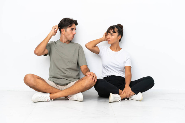 Young mixed race couple sitting on the floor isolated on white background having doubts while scratching head - Photo, Image