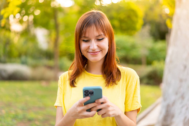 Dışarıdaki güzel kızıl saçlı kadın cep telefonuyla mesaj ya da e-posta yolluyor. - Fotoğraf, Görsel