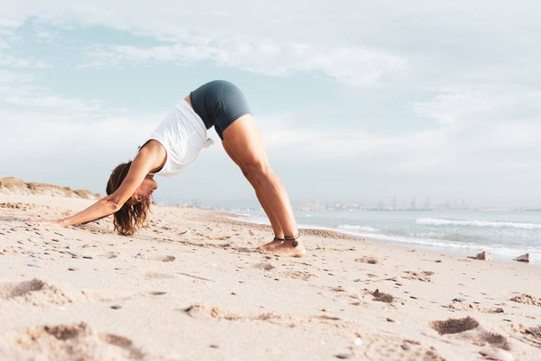 Woman on the beach doing the yoga posture adho mukha svanasana, with the sea at background - Photo, Image