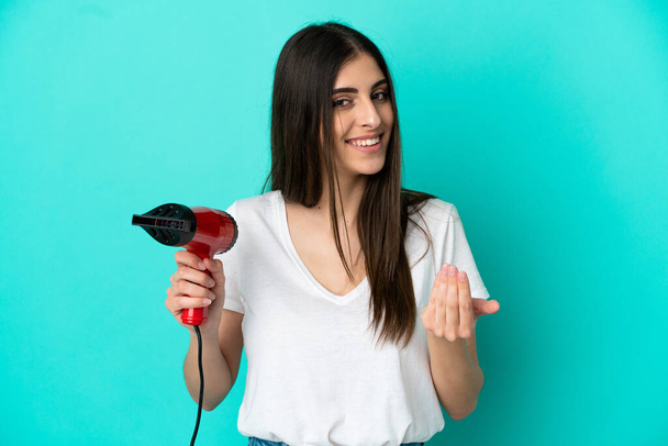 Young caucasian woman holding a hairdryer isolated on blue background inviting to come with hand. Happy that you came - Photo, Image