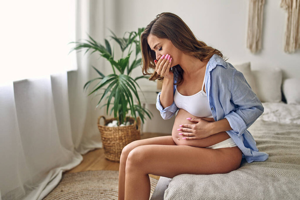 Mujer embarazada joven se enfermó sentada en la cama en el dormitorio sosteniendo su estómago y tosiendo. Malestar durante el embarazo. Concepto de maternidad. - Foto, Imagen
