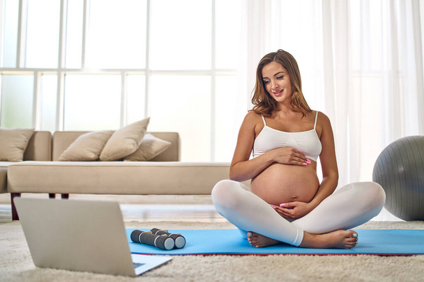 Young pregnant woman in sportswear doing exercises on a rubber mat in the living room with a laptop on video lessons. Sports and a healthy lifestyle during pregnancy. - Foto, Imagen